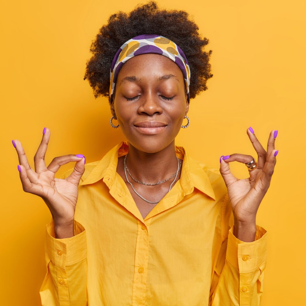 Relaxed peaceful dark skinned woman meditates with closed eyes practices yoga keeps hands in okay gesture wears casual shirt poses against vivid yellow background. Consideration and praying.